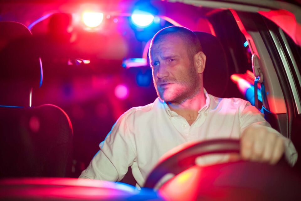Driver in car illuminated by police lights, showing a tense expression
